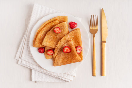 Top view of homemade traditional French folded crepe or blini sweet dessert snack decorated with sliced juicy strawberries served on round plate on napkin on white wooden tableの写真素材