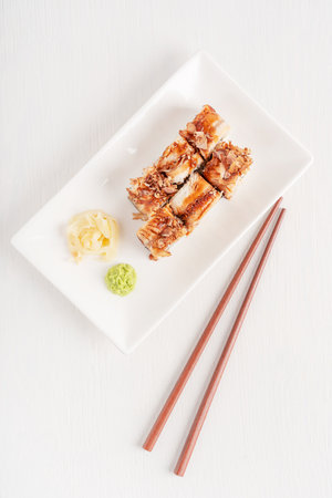 Top view of delicious homemade sushi rolls or unagi maki made from eel fish decorated with bonito flakes served on plate with ginger and wasabi on white wooden table with chopstickの写真素材