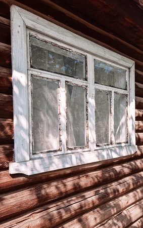 Old rural house or country hut facade wall made with weathered brown rough wooden planks with aged window with glass and white framing on sunny day with shadows in traditionalの写真素材