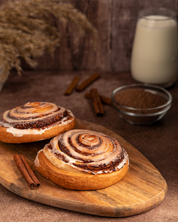Two tasty homemade cinnamon rolls bun with sugar icing on wooden cutting board on dark brown table with glass of milk and ingredients served for morning breakfast at rural cafeの写真素材