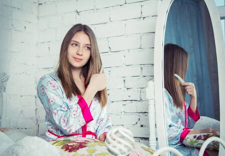 Beautiful blond teen girl combing her long hair on the bed near the mirrorの写真素材