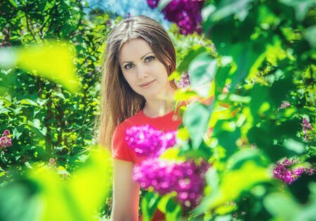 Beautiful blond girl in red dress looking from behind the bush of blooming lilac springtime outdoors