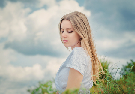 sad and pensive young woman sitting outdoors, clouded skyの写真素材