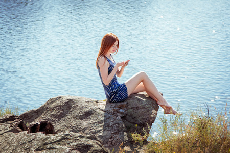 Pretty young redhaired woman in short dress sitting on the cliff edge above the river enjoying warm sunny summer day and looking at something on her handの写真素材