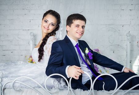 Attractive smiling happy bride and groom in ceremonial clothing sitting on a bedの写真素材