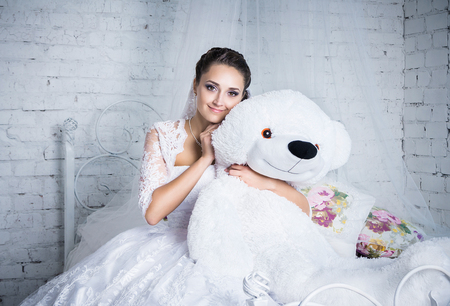 Pretty young brunette bride in a white wedding dress sitting on a bed embracing big white teddy bearの写真素材