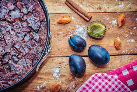 Chocolate clafoutis with plums in a baking pan, ripe fresh  plums and it`s stones, cinnamon, flour and cocoa powder on wooden backgroundの写真素材