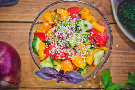 Fresh mixed vegetable salad with sesame in a glass bowl and ingredients - tomatoes, cucumbers, yellow pepper, red onion, basil and parsleyの写真素材