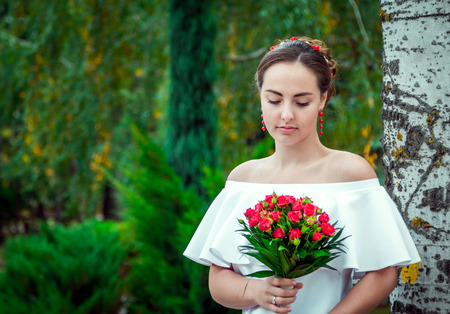 Portrait of beautiful young bride in white frilly dress with open shoulders and red earrings looking at wedding bouquet of small red roses in her hand, standing in the autumn parkの写真素材
