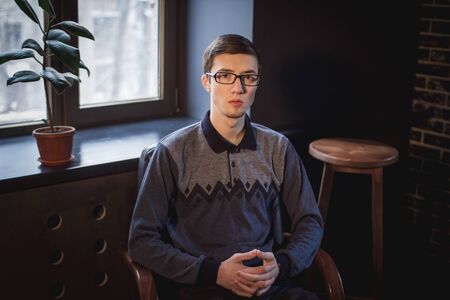 Portrait of handsome serious young man, wearing glasses, sitting in a dark room near the windowの写真素材