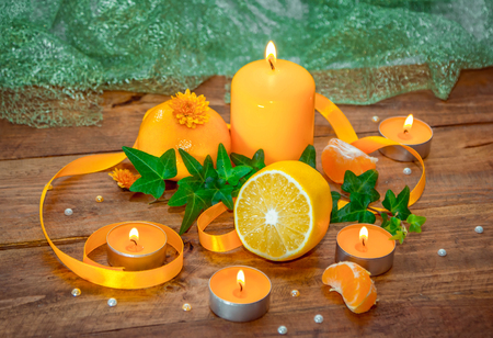 Citrus and herbs aroma concept. Burning yellow aroma candles, tangerine, lemon and ivy branch with green leaves, decorated by yellow satin ribbon, on a wooden backgroundの写真素材