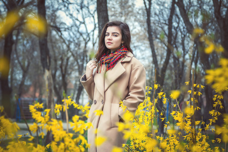 Beautiful teen girl, wearing trendy oversize beige coat and colorful scarf, walking in a spring park near blooming bushes with yellow flowersの写真素材