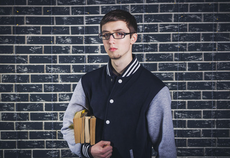 Studio portrait of serious male student. Young man, wearing glasses, holding books in front of dark brick wall.の写真素材