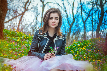 Beautiful teen girl wearing black leather jacket and pink tutu tulle skirt sitting on the spring meadow with yellow and purple flowers in the forestの写真素材