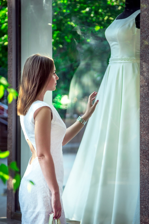 Elegant young woman admiring beautiful white wedding dress at the window of bridal boutique. Dreaming about wedding concept.の写真素材