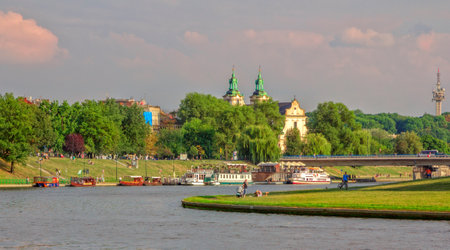 Krakow / Poland- August 14, 2017: citizens and tourists walking and relaxing on green lawns near Vistula river with view of boats, bridge and Catholic church of saint Bernadyna. Sunset skyのeditorial素材