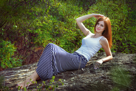 Beautiful young smiling woman with long red hair, wearing long striped skirt and white top, laying on a stone cliff in a forestの写真素材