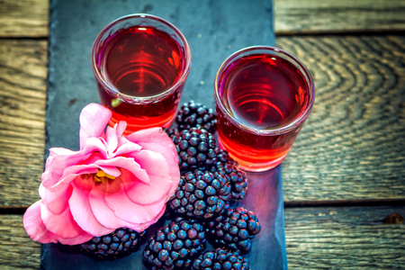 Homemade berry liqueur with fresh ripe blackberries, pink flower and black stone tray on a wooden background. Sweet alcohol drink.の写真素材