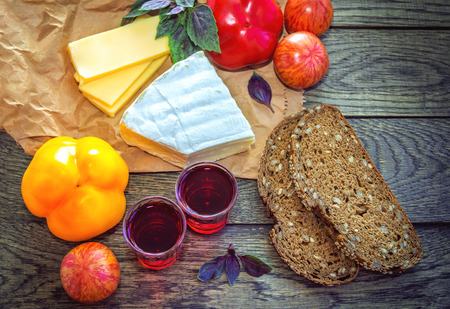 Served snack. Vegetables - tomatoes, red and yellow pepper, cheese, bread with sunflower seeds and sesame and homemade berry liqueur in shot glasses on a wooden tableの写真素材