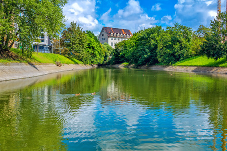 Summer cityscape - river surface with blue sky and green trees reflection, swimming duck and old...