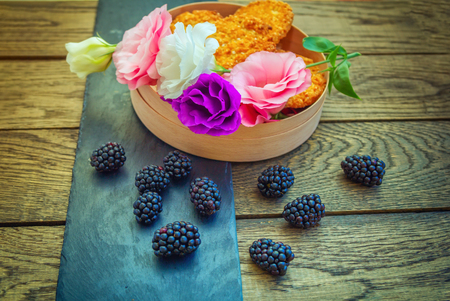 Fresh ripe blackberries, colorful eustoma flowers and biscuits in a box and black stone tray on a wooden background.の写真素材