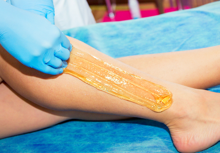 Close-up hands of cosmetologist in blue gloves applying paste for sugaring depilation, hair removal...