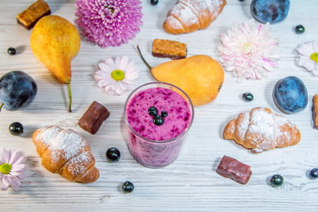 Berry smoothie in a glass, ripe fruits - pears and plums, flowers, croissants and chocolate candies on a wooden table surface. Sweet breakfast or snack.の写真素材