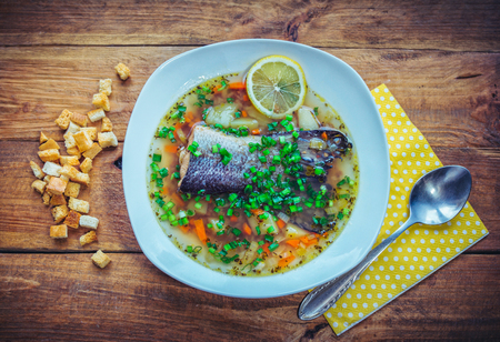 Fish soup with salmon tail, lemon slice and cut green onion in a plate with croutons and spoon on the wooden tableの写真素材