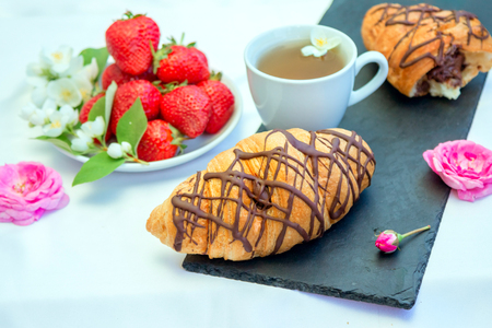 Herbal tea, beautiful pink rose flowers, ripe strawberry and chocolate croissant on the table. Sweet snack.の写真素材