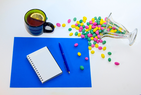 Blank spiral notepad, pen, cup of tea with lemon slice and glass vase with colorful candies on the table.の写真素材
