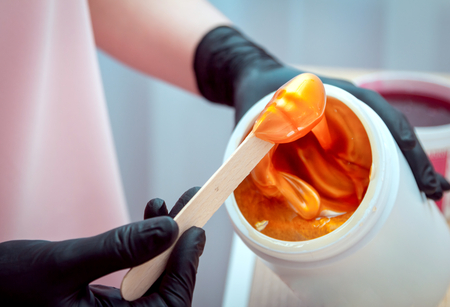 Close-up hands of cosmetologist in black gloves preparing golden colored waxing paste on spatula for sugaring depilation, hair removal beauty procedure.の写真素材