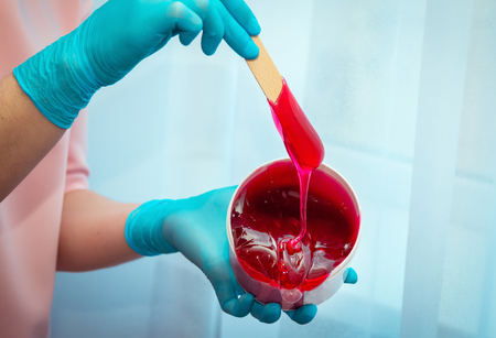 Close-up hands of cosmetologist in black gloves preparing red  berry flavor waxing paste on spatula for sugaring depilation, hair removal beauty procedure.の写真素材