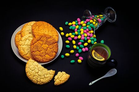 Homemade biscuits on a plate, glass vase with colorful candies and cup of tea with lemon slice on a black background. Sweet breakfast or snack.の写真素材