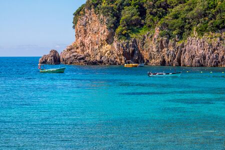 Beautiful landscape with sea and small colorful boats on a water surface, mountain and cliffs in a water, green trees. Corfu Island, Greece.の写真素材