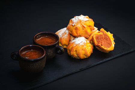 Fresh hot coffee in ceramic cups and sweet dessert â profiterole or eclair cakes with caramel cream and sugar powder on dark black backgroundの写真素材