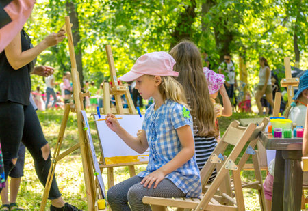 Zaporizhia / Ukraine-June 1, 2019: children participating at art and craft outdoors workshop, painting pictures with colorful gouache paints, sitting by wooden easel in a park on family charity festivalのeditorial素材