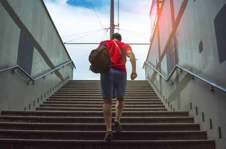 Man, wearing red t-shirt and jeans shorts, with black backpack, walking upstairs from subway....