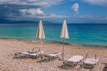 View of empty beach - white deck chairs and umbrellas near sea water, pebbles and golden sand, mountains on the horizon and dark clouds on the sky. Landscape of abandoned summer resort.の写真素材