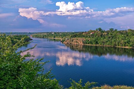 Beautiful spring landscape with river, lush forest, cliffs and white clouds, reflecting on calm blue water.の写真素材
