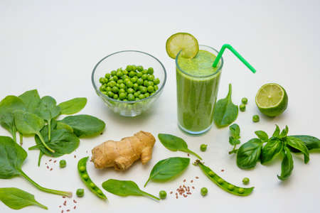 Smoothie in a glass and ingredients - green peas, ginger, basil and spinach leaves on a white background. Healthy vegan cocktail.の写真素材