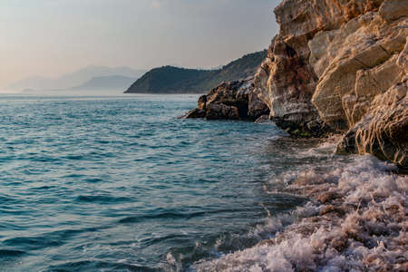 Beautiful landscape - beach in sunset sunlight - waving sea water, rocks and cliffs, blue sky and mountains on the horizon.の写真素材