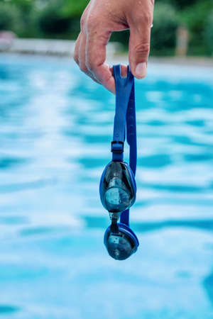 Man`s hand holding professional swimming goggles with pool blue water on backgroundの写真素材