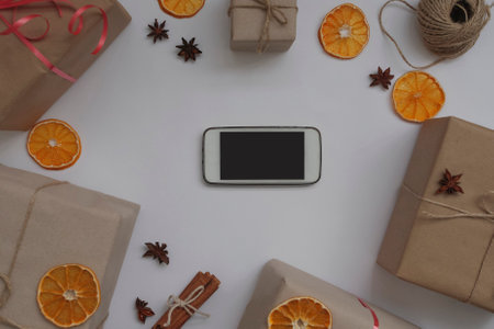 Christmas background with a phone with an empty space for advertising, gifts in craft paper, cinnamon, oranges, star anise on a white background. Advertising space, top view, flat layの写真素材
