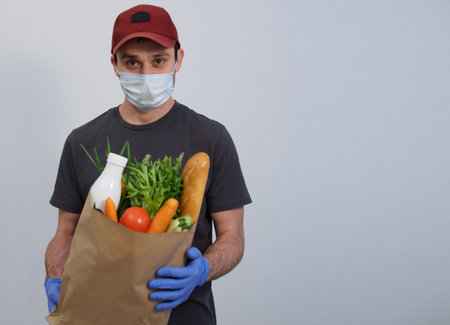 A home delivery worker wearing gloves and a mask holds a kraft paper bag with fresh food. Fresh vegetables, herbs, milk, bread in a package on a white background. Space for text, advertisements, announcements. Delivery of products at home for security reasons from COVID-19の写真素材