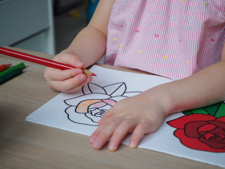 A girl in a pink striped T-shirt sits at a wooden table and draws a rose according to the pattern with a red pencil. Hands of a drawing girl close up. A 4-5 year old girl draws at home in quarantine. Creative development conceptの写真素材