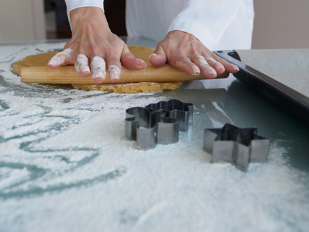 Close-up female hands roll out dough for cookies with a wooden rolling pin in the kitchen at home. New Year's metal forms for cookies in the shape of a Christmas tree and a star lie on the kitchen table. Mom prepares cookies for breakfastの写真素材
