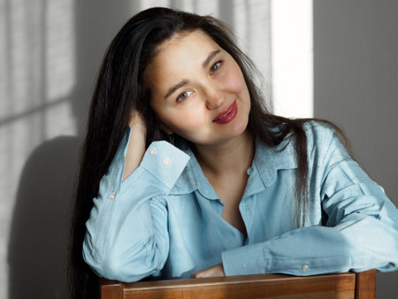 Young beautiful woman with long dark hair, expressive brown eyes sits on a chair by the window in a mens blue shirt, looks directly into the cameraの写真素材