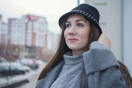 Portrait of a beautiful young woman enjoying a walk in the autumn city. A girl with long dark hair, a gray coat and a black hat looks to the side in the city centerの写真素材