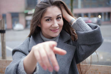 Portrait of a smiling brunette girl with long hair in a gray coat outside. Pretty girl points her finger directly at the camera and smiles, selective focus. Walk in the autumn cityの写真素材