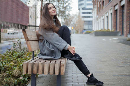 A young attractive girl in stylish clothes sits on a bench in the city center. A girl in a long gray coat and black jeans walks through the city streets in autumnの写真素材